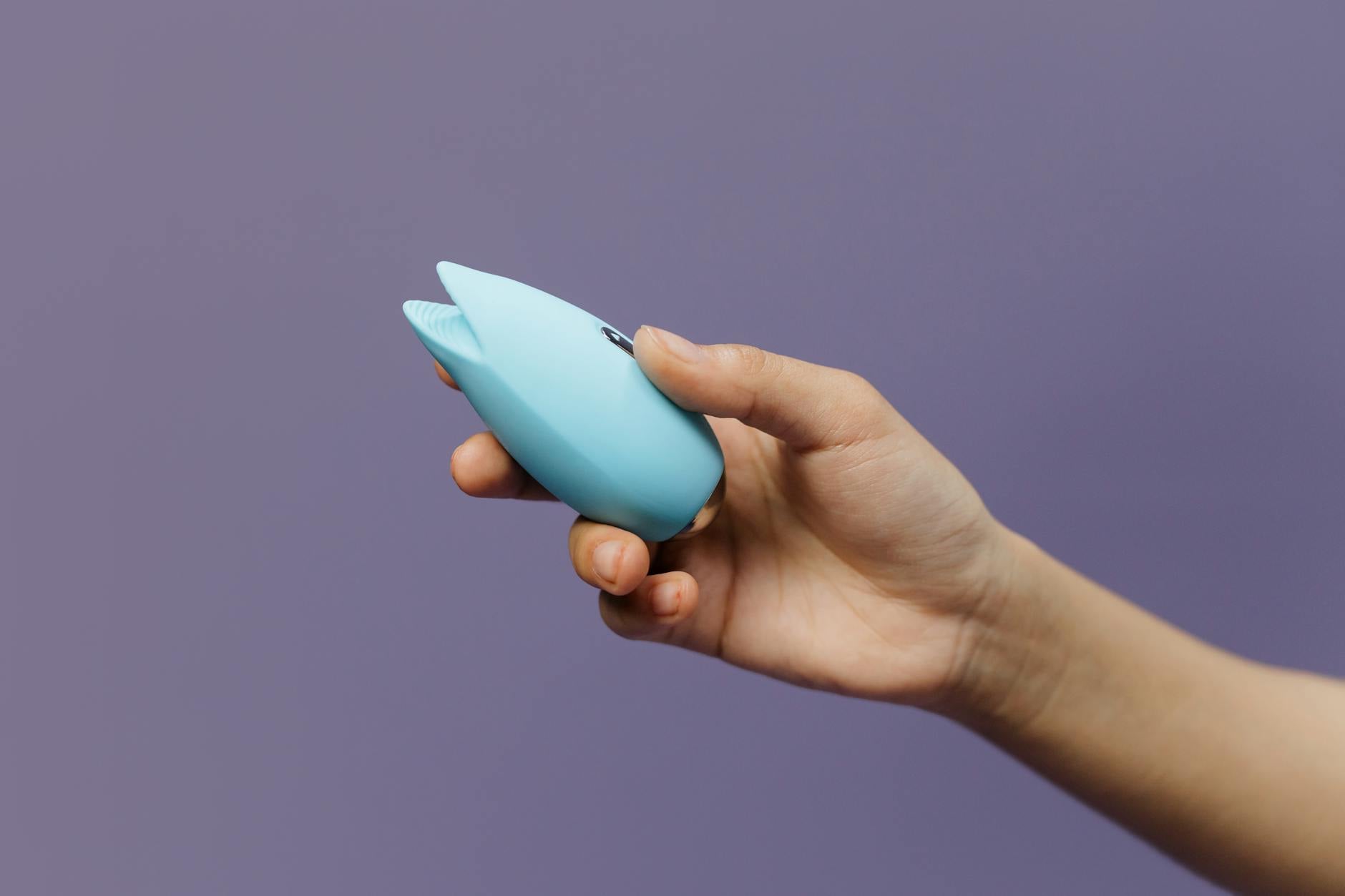 A blue silicone clitoral vibrator held gently in hand against a solid purple background